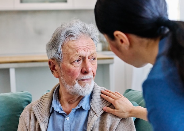 An older man with a white beard looks concerned while a woman gently rests her hand on his shoulder, offering comfort in a home setting.