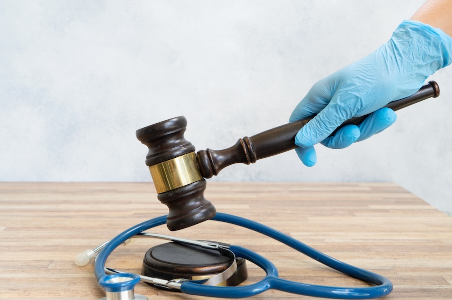 A hand in a blue medical glove holds a judges gavel above a stethoscope on a wooden surface, symbolizing medical law or healthcare justice.