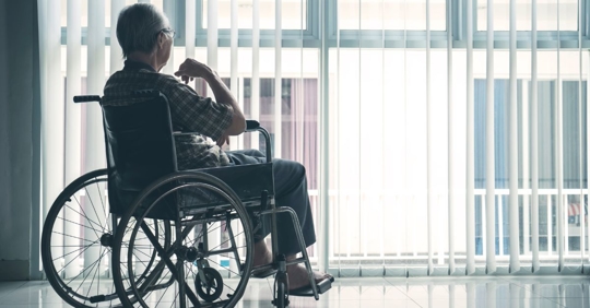 An elderly person in a wheelchair sits indoors, looking out a large window with vertical blinds. Soft daylight filters into the room, creating a calm, reflective atmosphere.