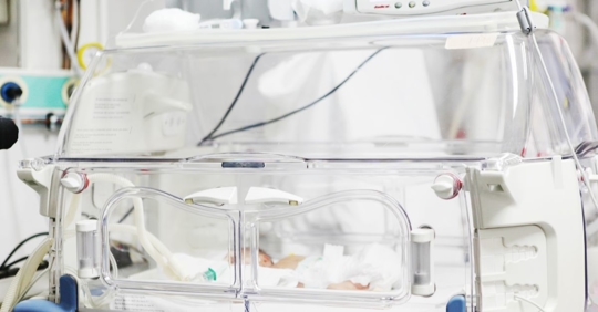 A hospital incubator with a newborn baby inside, surrounded by medical equipment and tubes, in a neonatal intensive care unit.
