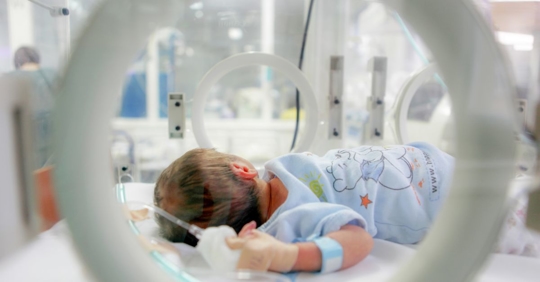 A newborn baby lies in an incubator in a hospital, wearing a hospital gown and identification band, with medical equipment visible in the background.
